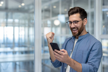 Young man in blue shirt standing in a modern office, expressing joy and excitement while looking at his smartphone, celebrating a significant achievement or good news