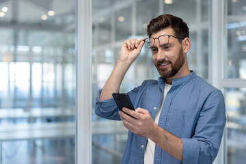 Entrepreneur man lifting eyeglasses from his face, focusing on the smartphone screen while checking messages or browsing on his mobile device in a modern office environment