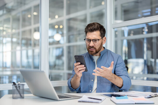 Upset businessman is experiencing anger and stress while dealing with a problem on his mobile phone in a contemporary glass-walled office environment, expressing strong negative emotions