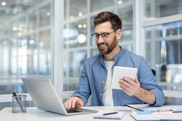 Young man in glasses and casual shirt smiling, multitasking with a laptop and digital tablet, sitting at a white desk in a modern brightly lit office, engaging with technology