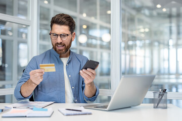 Man at a desk in a modern office is looking frustrated and upset while holding a credit card and...