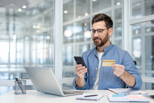 Happy man making a purchase on his mobile phone with a golden credit card in hand, sitting at a desk with a laptop, enjoying convenient e-commerce and secure digital payment
