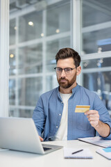 Man holding credit card while using laptop in a modern office, making secure online payments and...