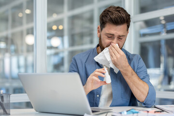 Young businessman experiencing symptoms of cold, flu, or seasonal allergy while working on a laptop, using a tissue and holding a nasal spray bottle in a modern office environment