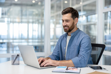 Young man smiling and typing on a laptop, concentrating while working at his desk in a bright, contemporary open-plan office setting, representing productivity and a positive work environment