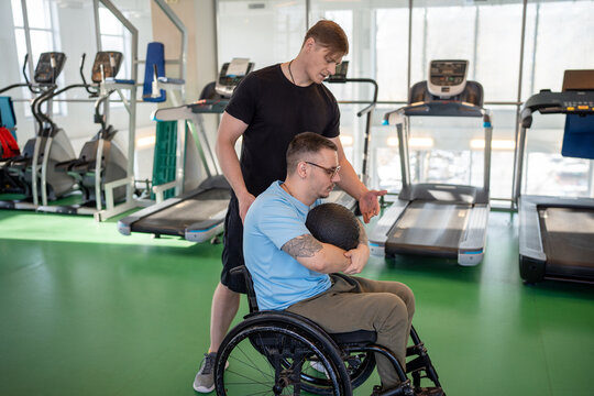 Determined man in wheelchair hold weighted medicine ball to rotate trunk and lift with two hands. Trainer guide movement to activate core and upper body, inclusive fitness, functional rehabilitation