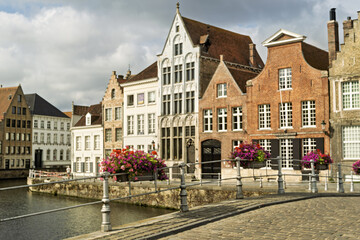 Fototapeta premium Row of traditional buildings along a canal in Bruges, Belgium, featuring stepped gables and facades in beige, white, and brick red. The calm canal reflects the architecture,