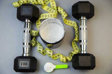 Black dumbbells are positioned alongside a measuring tape and a protein powder container, showcasing fitness essentials for a workout routine and healthy living