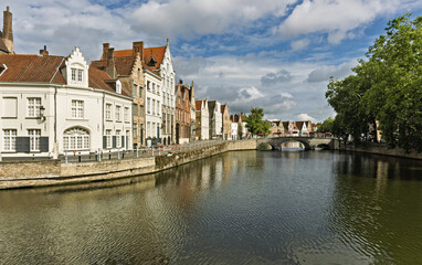 Obraz premium Row of traditional buildings along a canal in Bruges, Belgium, featuring stepped gables and facades in beige, white, and brick red. The calm canal reflects the architecture,