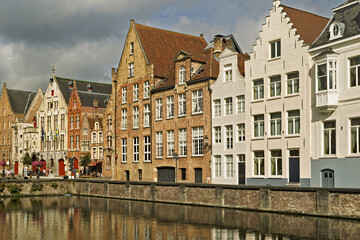 Row of traditional buildings along a canal in Bruges, Belgium, featuring stepped gables and facades in beige, white, and brick red. The calm canal reflects the architecture,