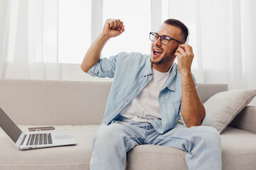 Fototapeta premium Energetic man enjoying music on headphones, sitting on a modern sofa with a laptop, celebrating his creative break. Perfect for showcasing relaxation and productivity.