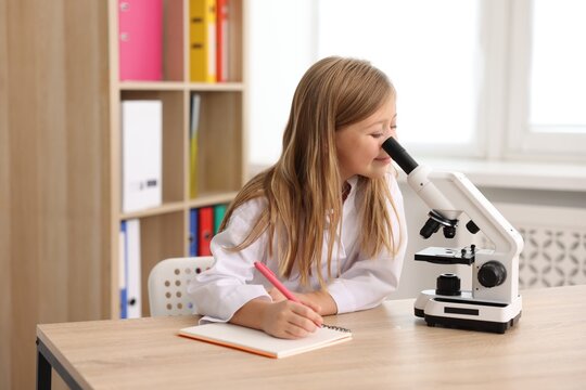 Cute little girl with pen, book and microscope at desk indoors. Child and science