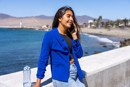 Young latin American woman talking on phone while relaxing by the coastline on a sunny day