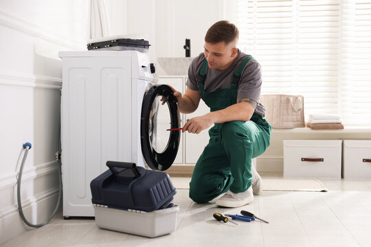 Repairman in uniform fixing broken washing machine with screwdriver at home