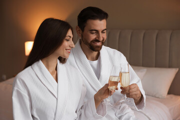 Happy couple in bathrobes with glasses of sparkling wine on bed indoors
