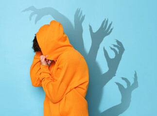 Scared teenage boy hiding on light blue background. Shadows of creepy hands behind him. Phobia,...
