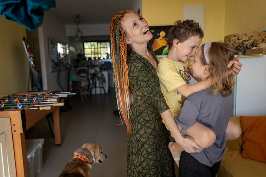 Joyful mother with bright orange and green dreadlocks laughs, embracing her smiling son and daughter tightly in a warm family hug at home, with a happy dog nearby.