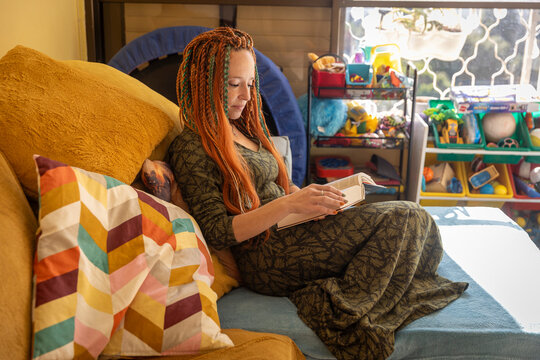 A woman with calm, focused expression and long dreadlocks sits relaxed on a sofa, deeply engrossed in reading a book in a sunlit corner of the room.