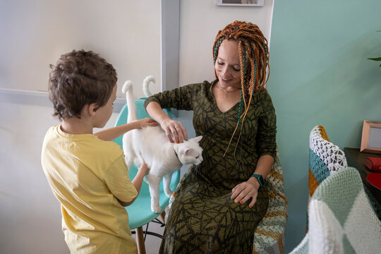 A cheerful mother with colorful dreadlocks and her son affectionately pet their white cat as they sit together, sharing a joyful moment.