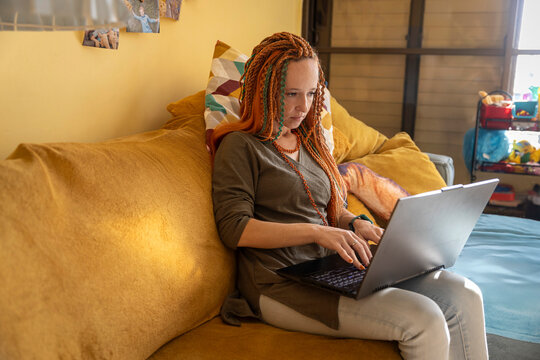 A diligent woman with long dreadlocks sits on a sofa, seriously concentrating on her work on a black laptop.