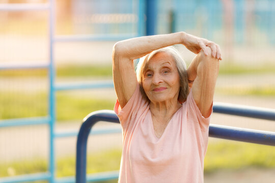 Elderly woman in a light pink top performs an overhead triceps stretch on a sunny day, smiling at the camera, embodying joy, fitness, and healthy, active aging.