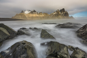View of jagged, snow-capped mountains rise majestically from the misty sea, framed by dark, rocky foreground, creating a serene yet dramatic landscape, Stokkness, Iceland.