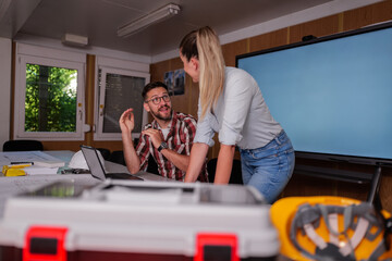 Two architects, a man and a woman, discussing blueprints and collaborating on a laptop in their office within a construction container, highlighting teamwork in design