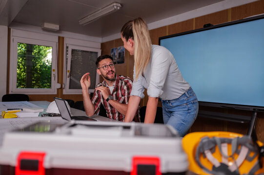 Two architects, a man and a woman, discussing blueprints and collaborating on a laptop in their office within a construction container, highlighting teamwork in design