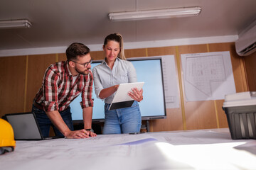 Two architects are examining blueprints and using a digital tablet during a meeting in their office. Collaborating on a construction project and utilizing technology for efficient design and planning