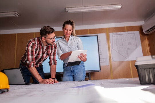 Two architects are examining blueprints and using a digital tablet during a meeting in their office. Collaborating on a construction project and utilizing technology for efficient design and planning - Powered by Adobe