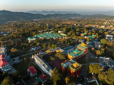 Aerial view of golden-roofed monastery complexes nestled amidst the lush green canopy of trees, contrasting with the distant blue mountains, Bir, Himachal Pradesh, India.