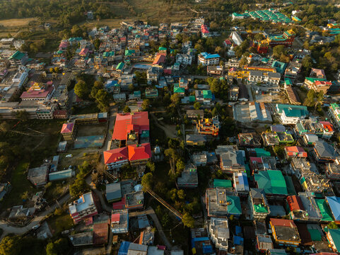 Aerial view of a vibrant town with buildings displaying varied roof colors, nestled amidst lush trees, casting long shadows in the golden sunlight, Bir, Himachal Pradesh, India.
