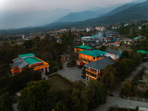 Aerial view of buildings with orange walls and green roofs nestle among the lush greenery against a backdrop of hazy mountains, Bir, Himachal Pradesh, India.