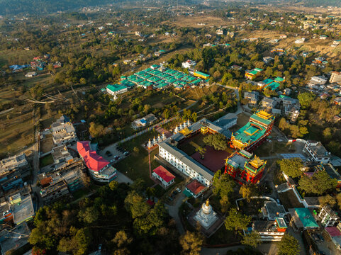 Aerial view of golden-roofed monasteries nestled amidst lush greenery and terracotta-roofed buildings, under soft sunlight, creating a serene tapestry of color, Bir, Himachal Pradesh, India.