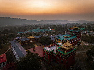 Aerial view of vivid red and gold monastery architecture, set against a backdrop of rolling hills and a soft, pastel sunrise, Bir, Himachal Pradesh, India.