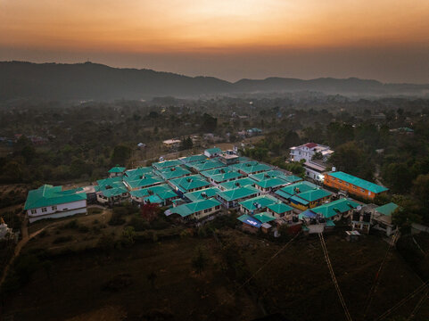 Aerial view of rows of buildings with green roofs nestled amidst lush trees, with mountains silhouetted against a warm sunset, Bir, Himachal Pradesh, India.