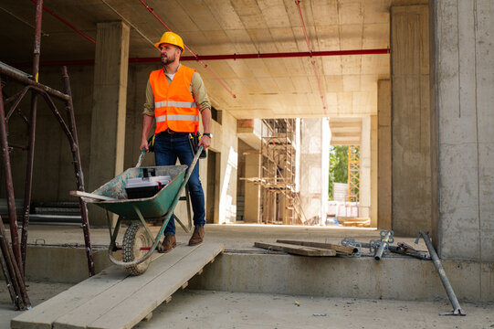 Construction worker wearing a safety vest and helmet, pushing a wheelbarrow loaded with a toolbox across a wooden plank at a bustling building site