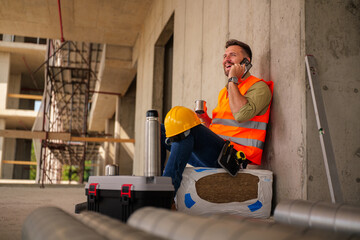Cheerful construction worker enjoying a coffee break while chatting on a mobile phone, sitting on insulation material with tools and safety helmet nearby