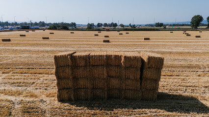 A large stack of rectangular straw bales sitting in a harvested field with stubble, overlooking a busy road and distant trees
