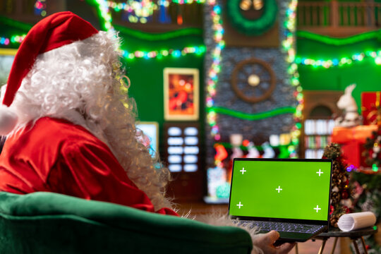 Santa Claus using mockup laptop in room with colorful garlands holiday props to check naughty and nice kids list. Santa in festive lights decorated workshop preparing gifts using green screen device