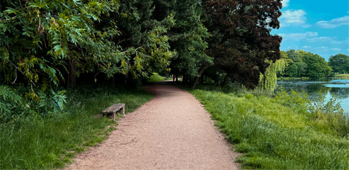 A serene path winds through lush greenery along the edge of a peaceful lake. A wooden bench offers a spot for rest and reflection in Roundhay Park, Leeds, UK
