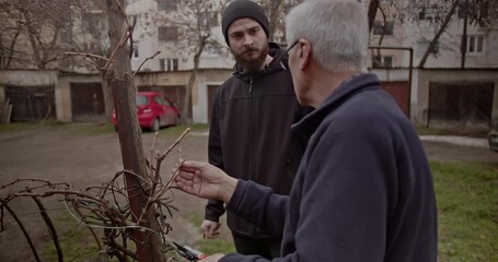 An experienced senior man demonstrates pruning techniques on a bare plant to a young, attentive learner. This scene highlights inter generational knowledge transfer and practical gardening skills.