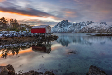 View of a red cabin standing on stilts in the water, reflecting the colorful sunrise sky, with snow-capped mountains in the background, Reine, Nordland, Norway.