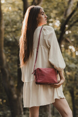 Summer Freedom and Style: A woman in a bright, airy dress with a red/burgundy crossbody bag, illuminated by the rays of the setting sun in the forest