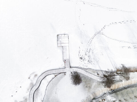 Aerial view of a snow-covered pier extending into the frozen lake, its stark white contrasting with the dark lines of footsteps across the icy expanse, Trakai, Trakai, Lithuania.