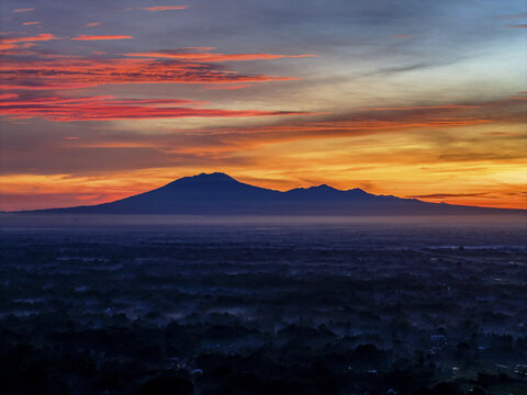 Aerial view of a majestic mountain range silhouetted against a vibrant sunrise sky, casting a warm glow over the landscape, Klaten Regency, Central Java, Indonesia.