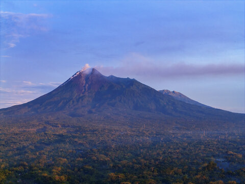 Aerial view of the majestic Mount Merapi looms over the lush green forest, its peak shrouded in a delicate veil of mist, Klaten Regency, Central Java, Indonesia.