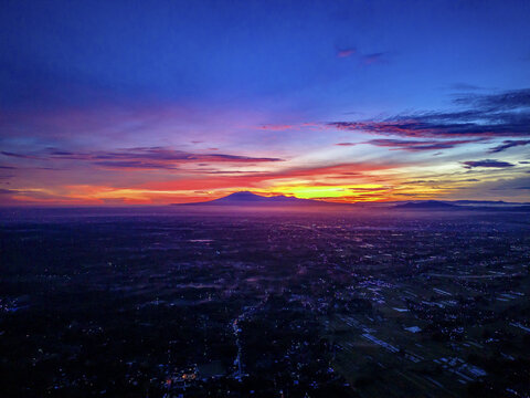 Aerial view of a vibrant sunrise casting a warm glow over the landscape near the distant silhouette of Mount Lawu, Klaten Regency, Central Java, Indonesia.