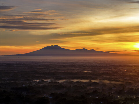 Aerial view of a distant lawu mountain range silhouetted against a vibrant sunrise with orange and yellow hues, contrasting with the dark landscape below, Klaten Regency, Central Java, Indonesia.