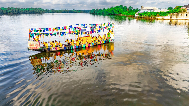 Aerial view of a vibrant boat adorned with colorful flags and passengers in yellow shirts sailing on the river, Buguma, Rivers State, Nigeria.